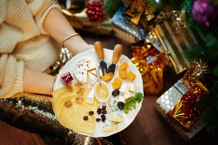 Closeup on trendy middle age woman in gold sequin skirt and white sweater under decorated Christmas tree near present boxes holding cheese platter.の写真素材