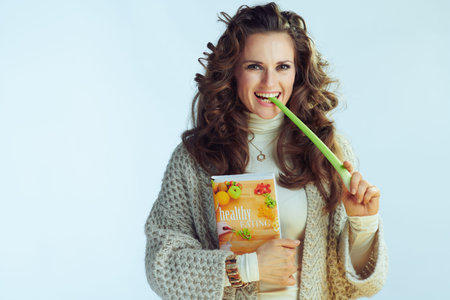 happy modern housewife with long wavy hair in neck sweater and cardigan with healthy eating book eating celery isolated on winter light blue background.の写真素材