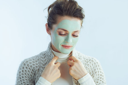 Portrait of pensive elegant housewife in roll neck sweater and cardigan with green facial mask on winter light blue background.の写真素材