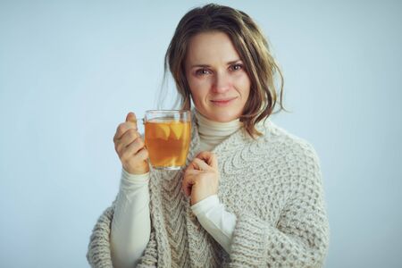 Portrait of sick modern 40 years old housewife in roll neck sweater and cardigan showing cup of hot tea with ginger, lemon and honey isolated on winter light blue background.の写真素材