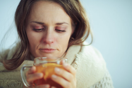Portrait of sad sick modern middle age woman in roll neck sweater and cardigan drinking cup of hot...