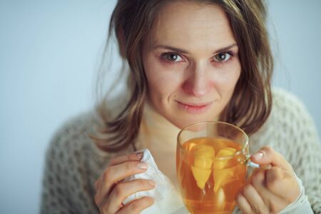 Portrait of ill elegant woman in roll neck sweater and cardigan drinking cup of hot tea with ginger, lemon and honey isolated on winter light blue background.の写真素材