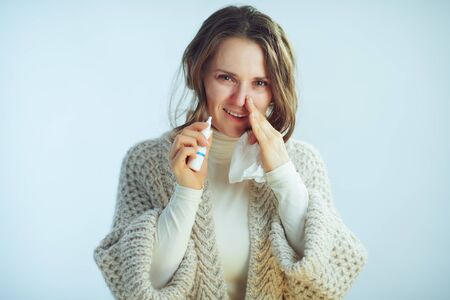 Portrait of ill modern middle age woman in roll neck sweater and cardigan with napkin using nasal spray isolated on winter light blue background.の写真素材