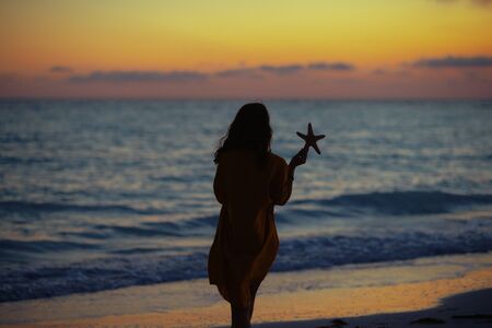 Silhouette of healthy woman with long brunette hair on the seashore at sunset holding starfish.の写真素材