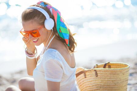 happy trendy woman in white t-shirt and pink shorts listening to the music with headphones on the ocean shore.