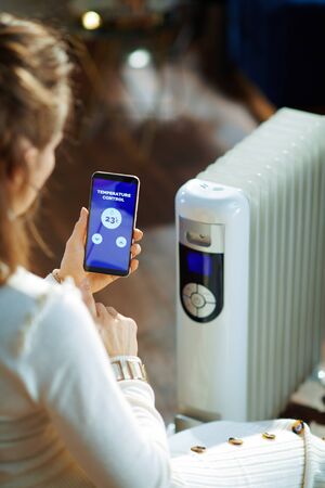 Trendy woman in white sweater and skirt in the modern living room in sunny winter day sitting near couch and white electric oil radiator using smartphone app for temperature control.の写真素材