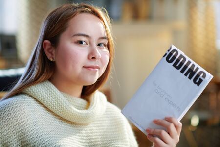 happy modern teenager girl with red hair in white sweater at modern home in sunny winter day holding educational coding book.の写真素材