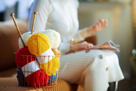 Closeup on basket with knitting yarn and needles and woman reading magazine in background in the...