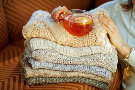 Closeup on elegant middle age woman in white sweater and skirt with pile of sweaters and cup of tea...
