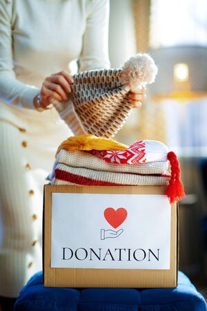 Closeup on trendy female in white sweater and skirt packing donation box with old warm clothes at modern home in sunny winter day.の写真素材