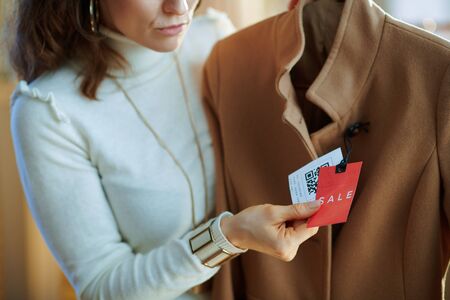 Closeup on concerned young woman in white sweater and skirt in modern fashion shop examines sale price tag on beige coat.の写真素材