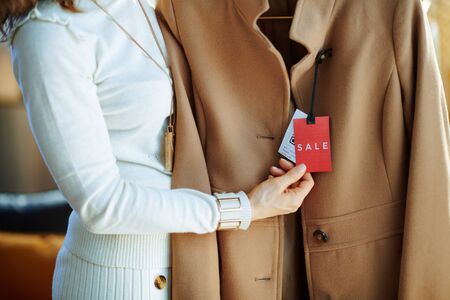 Closeup on woman in white sweater and skirt in modern fashion store examines sale price tag on beige coat.の写真素材