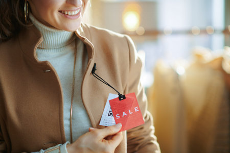 Closeup on young female in white sweater and skirt in modern fashion showroom trying beige coat and looking at red sale price tag.の写真素材