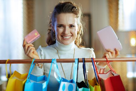smiling trendy middle age woman in white sweater and skirt with credit card and wallet near colorful shopping bags hanging on copper clothes rail.の写真素材