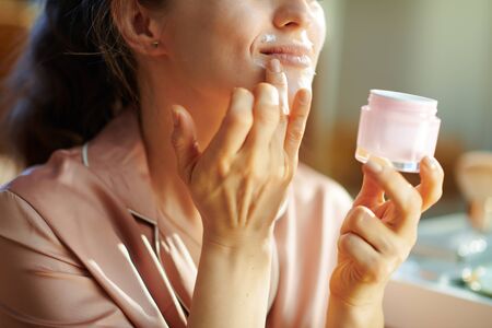 Closeup on happy modern woman in pajamas with pink cosmetic jar applying lip contour cream at modern home in sunny winter day.の写真素材