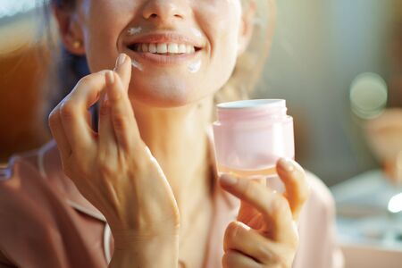 Closeup on happy elegant woman in pajamas with pink cosmetic jar applying lip contour cream in the modern house in sunny winter day.の写真素材