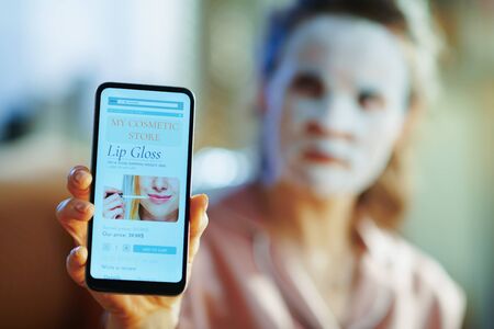 Closeup on elegant middle age woman in pajamas with white sheet facial mask on face showing smartphone with beauty makeup online shopping site at modern home in sunny winter day.の写真素材
