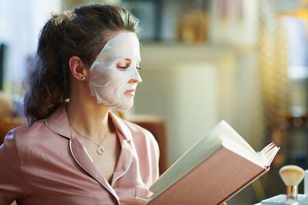 modern middle age housewife in pajamas with white sheet facial mask reading book at modern home in sunny winter day.の写真素材