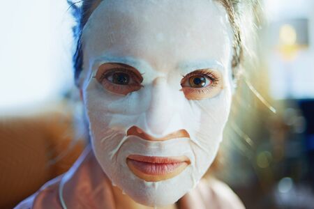 Portrait of elegant 40 years old woman in pajamas with white sheet facial mask on face at modern home in sunny winter day.の写真素材