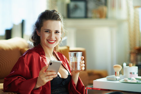 happy elegant female in black body lingerie and red bathrobe with cup of water near table with toiletries using water tracking app on a smartphone in the modern living room in sunny day.の写真素材