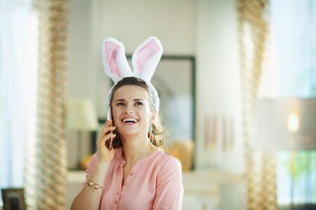 smiling elegant middle age housewife in a pink blouse and easter bunny ears in the modern living room in sunny spring day using a mobile phone.の写真素材