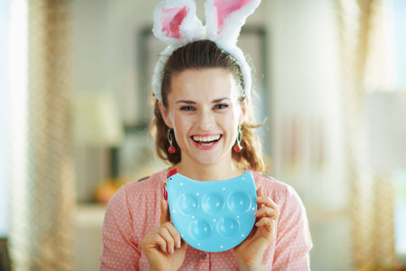 Portrait of smiling young woman in a pink blouse and easter bunny ears at modern home in sunny...