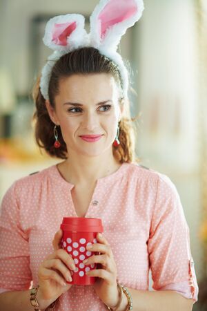 pensive modern woman in a pink blouse and easter bunny ears in the modern living room in sunny spring day holding coffee cup.の写真素材