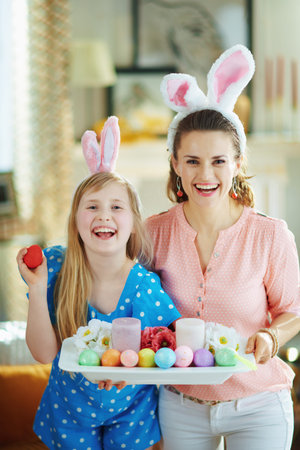 smiling elegant mother and daughter in easter bunny ears showing easter decorated plate with...