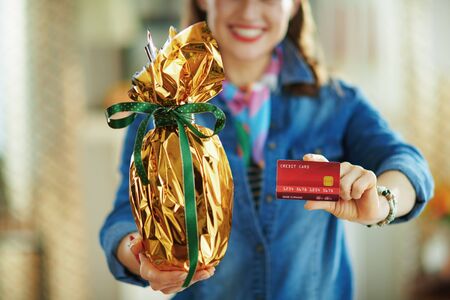 Closeup on smiling woman in the house in sunny spring day showing credit card and holding wrapped in gold foil big easter egg.の写真素材