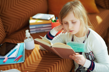 young child in white striped t-shirt with medical mask sitting near sofa and reading book in the...