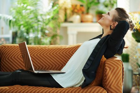 relaxed trendy 40 years old woman in white blouse and black jacket with laptop sitting on sofa in the modern living room in sunny day.の写真素材