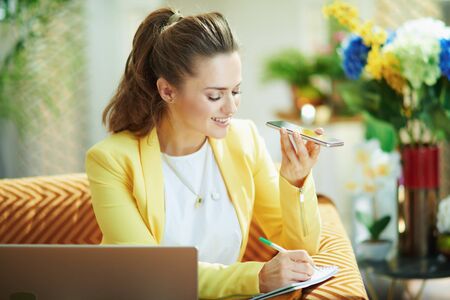 smiling stylish housewife in jeans and yellow jacket with laptop and smartphone study online at modern home in sunny day.の写真素材