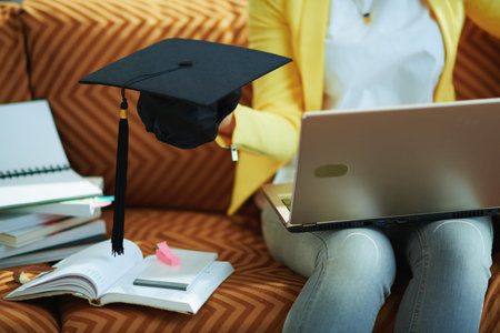 happy trendy middle age housewife in jeans and yellow jacket with laptop showing graduation cap at...