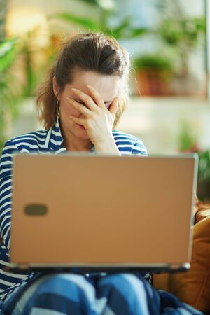stressed trendy woman in blue blouse and striped jacket sitting on sofa with laptop in the modern living room in sunny day.の写真素材