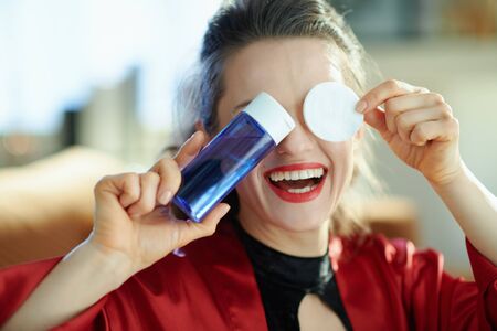smiling stylish woman in black body lingerie and red bathrobe showing makeup remover with cotton pads in the modern living room in sunny day.の写真素材
