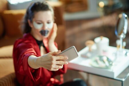 Closeup on female in black body lingerie and red bathrobe with black hydrogel lip mask and eye patches taking selfie with smartphone in the living room in sunny day.の写真素材