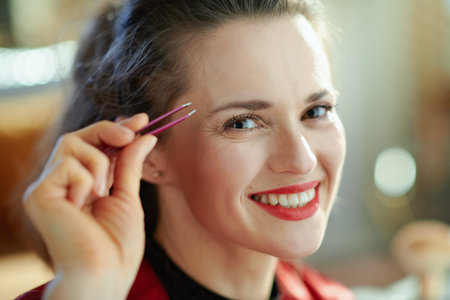 Portrait of happy modern woman in black body lingerie and red bathrobe plucking eyebrows with...