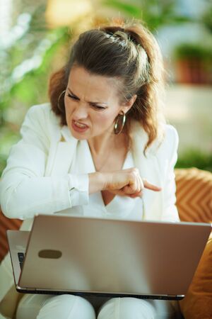 mad modern female in white blouse and jacket sitting on couch with laptop at modern home in sunny day.の写真素材