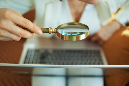 Closeup on woman in white blouse and jacket with laptop using magnifying glass at home in sunny day.