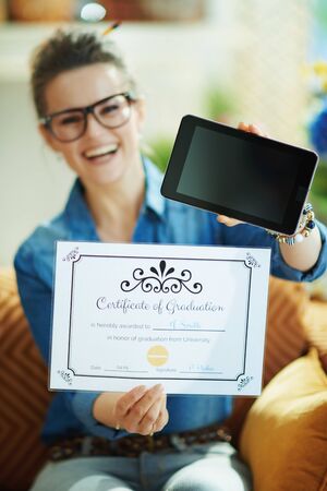 Closeup on smiling woman in jeans shirt in the living room in sunny day showing Certificate of Graduation and tablet PC blank screen.の写真素材