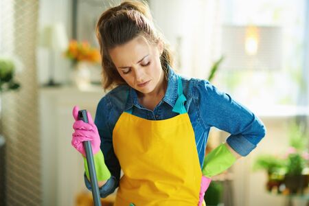 middle age woman in orange apron and pink rubber gloves with mop having back pain in the modern living room in sunny day.の写真素材