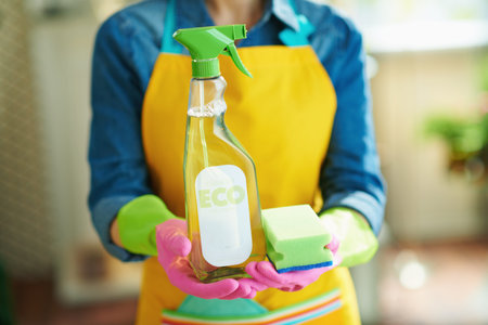 Closeup on woman in orange apron and pink rubber gloves with spray bottle of green cleaning supplies...