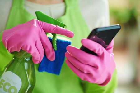 Closeup on woman in green apron and pink rubber gloves with spray bottle of green cleaning supplies and sponge using home cleaning app on smartphone at home in sunny day.の写真素材