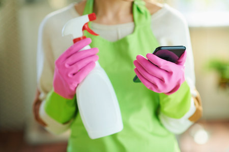 Young woman in green apron and pink rubber gloves with spray bottle of cleaning supplies reading about cleaning product on Internet using smartphone at modern home in sunny day.の写真素材