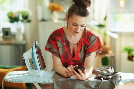 trendy middle age woman in red blouse and grey pencil skirt with iron checking how to iron correctly on internet on a smartphone in the modern living room in sunny day.の写真素材