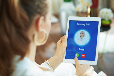 Seen from behind woman at home in sunny day taking incoming video call from doctor using telehealth...