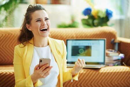 happy elegant middle age woman in yellow jacket with laptop and smartphone rejoicing while study online in the modern house in sunny day.の写真素材
