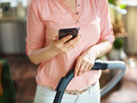 Closeup on woman in blouse and white pants with vacuum cleaner brush using home cleaning app on smartphone in the house in sunny day.の写真素材