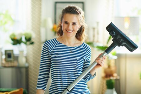 Portrait of happy young woman in striped t-shirt and white pants with vacuum cleaner in the modern living room in sunny day.の写真素材