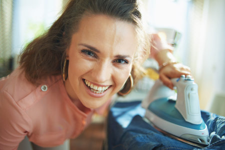 Portrait of happy young housewife in pink shirt and white pants at modern home in sunny day ironing on ironing board.の写真素材